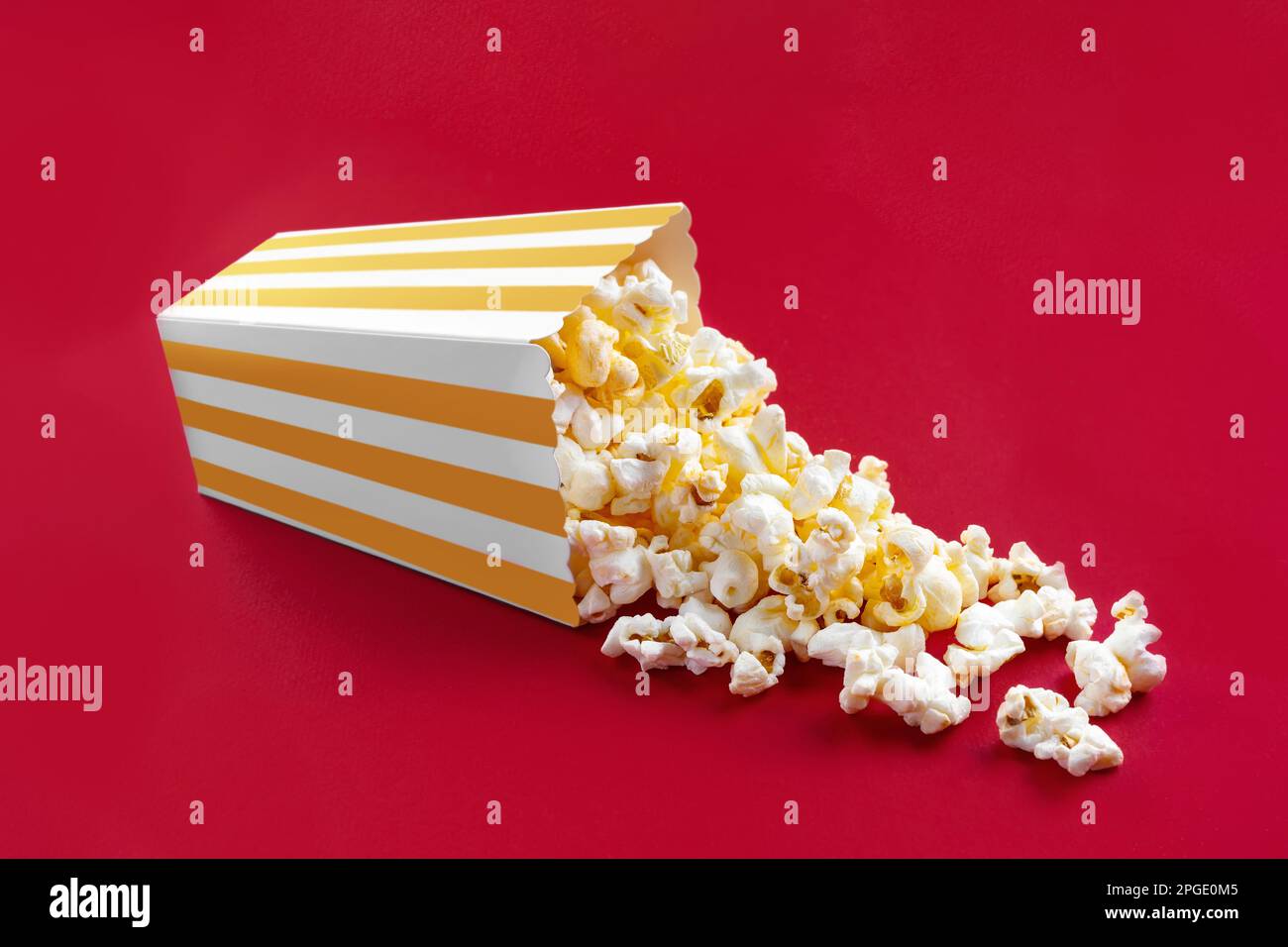 Gustoso popcorn al formaggio che cade da un secchio di cartone a strisce gialle, isolato su sfondo rosso. Dispersione di grani di popcorn. Film, cinema e ingresso Foto Stock