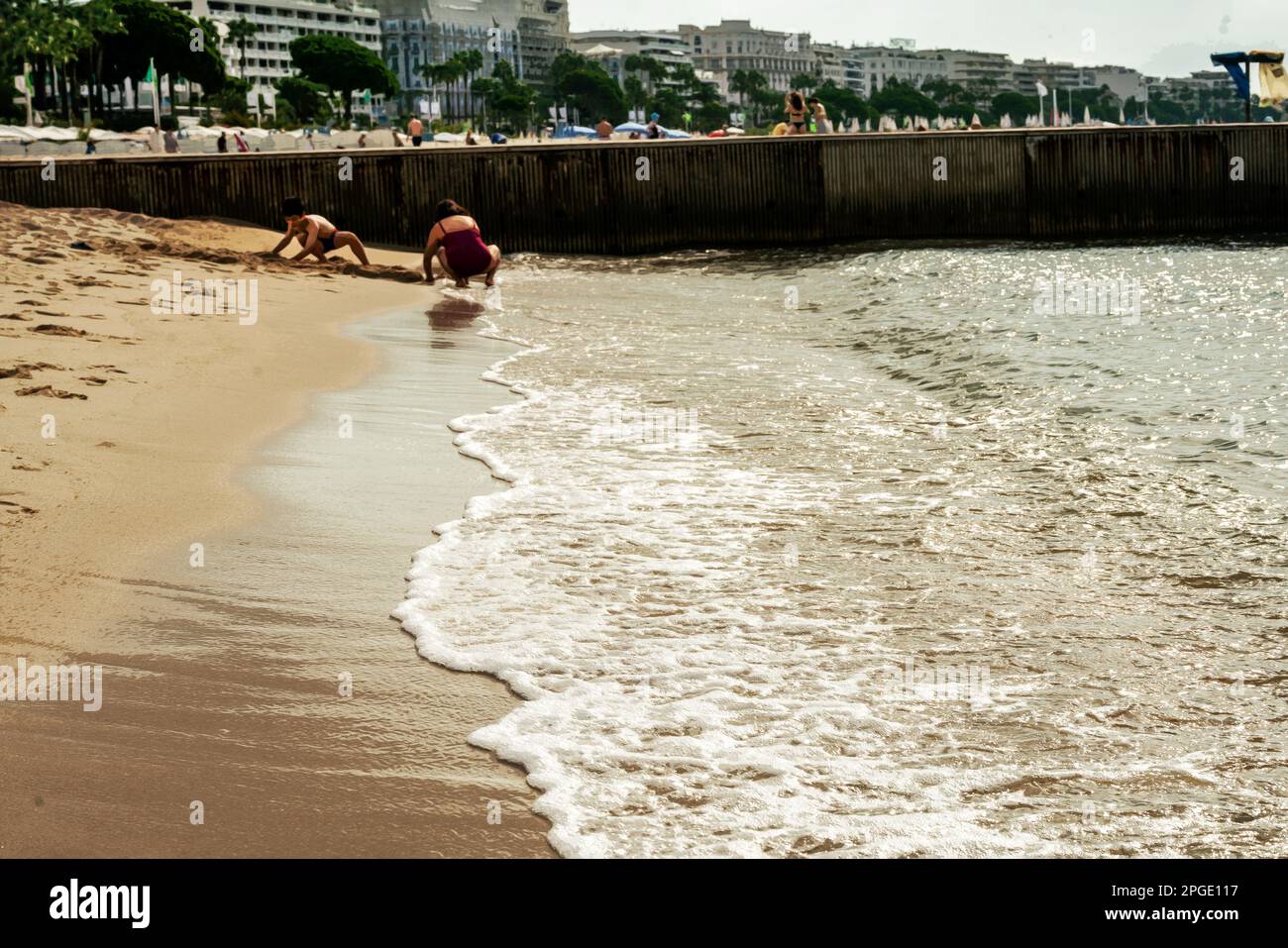 Un'onda di calma di una spiaggia pubblica rotola sulla sabbia. Madre e figlio (gente irriconoscibile) costruendo un castello di sabbia a Cannes. Croisette con palma tr Foto Stock