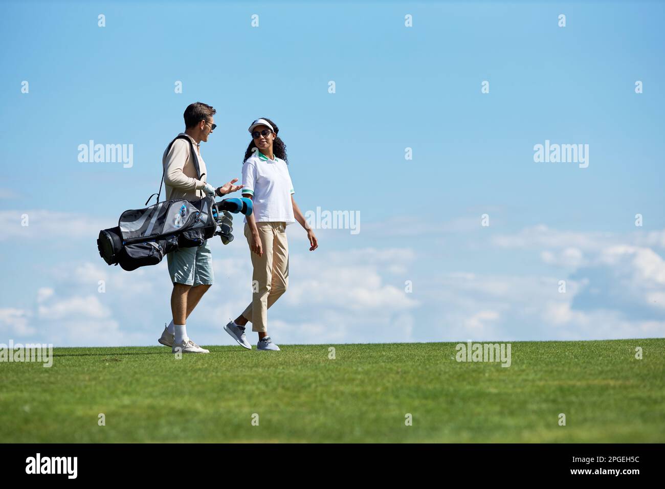 Ritratto con vista laterale minimale di un'elegante coppia sportiva che trasporta una borsa da golf che cammina sul campo verde contro il cielo blu, spazio copia Foto Stock