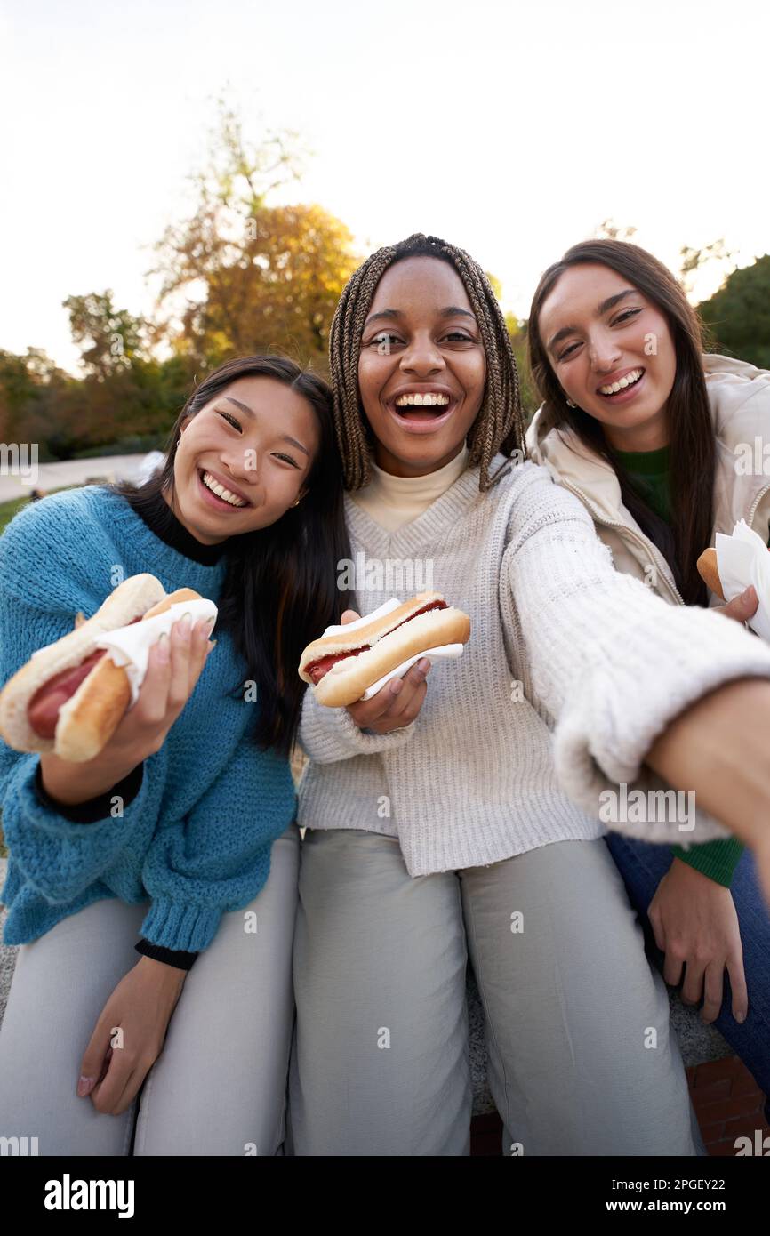 Gruppo verticale ragazze allegre selfie mangiare cibo di strada da asporto seduti sulla panca nella bella zona della città. Foto Stock