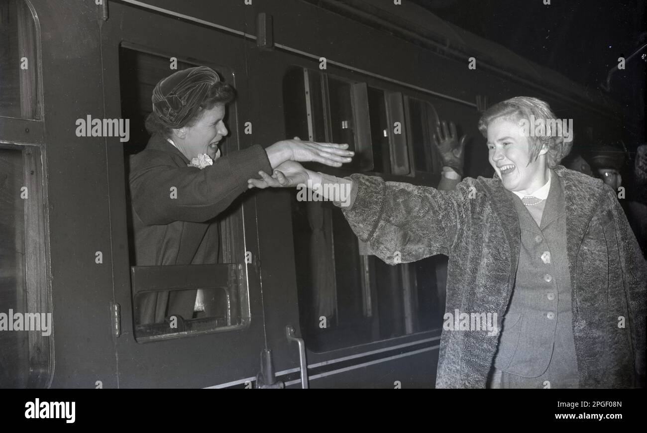 Intorno agli anni '1940, storica, la mano dell'amicizia, del tempo serale e una donna felice su un treno si appoggia fuori da una finestra aperta della carrozza per salutare la sua amica sorridente sulla piattaforma, che ha il braccio allungato per salutarla, Eugene, Oregon, USA. Foto Stock