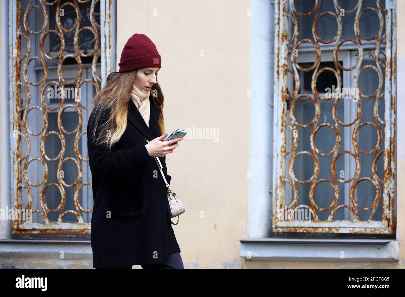 Giovane donna in cappotto a piedi con smartphone su una strada sullo sfondo di un muro di costruzione con vecchie finestre. Uso del telefono cellulare nella città primaverile Foto Stock