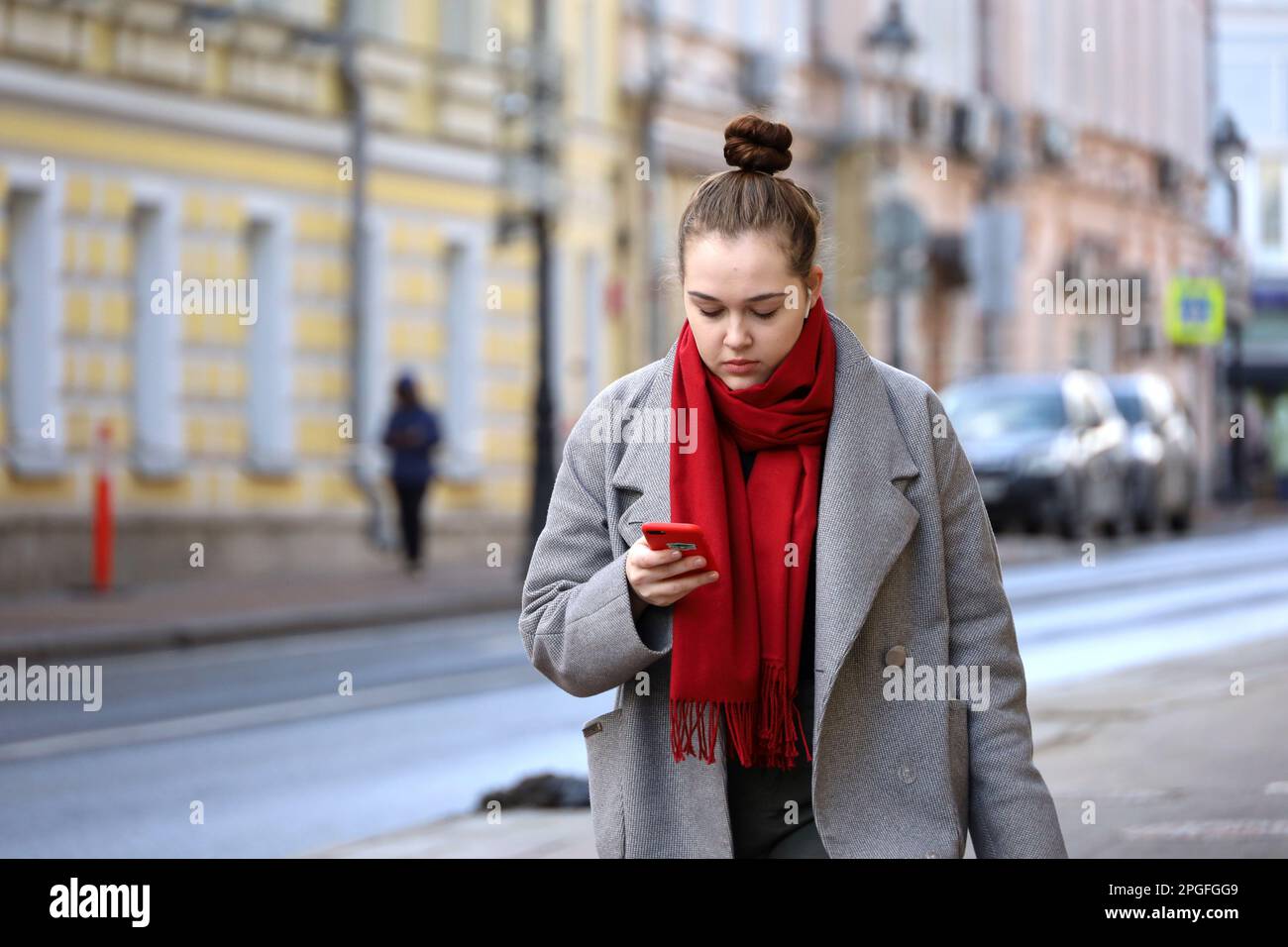 Giovane donna in cappotto e sciarpa rossa che cammina con lo smartphone su una strada. Uso del telefono cellulare nella città primaverile Foto Stock