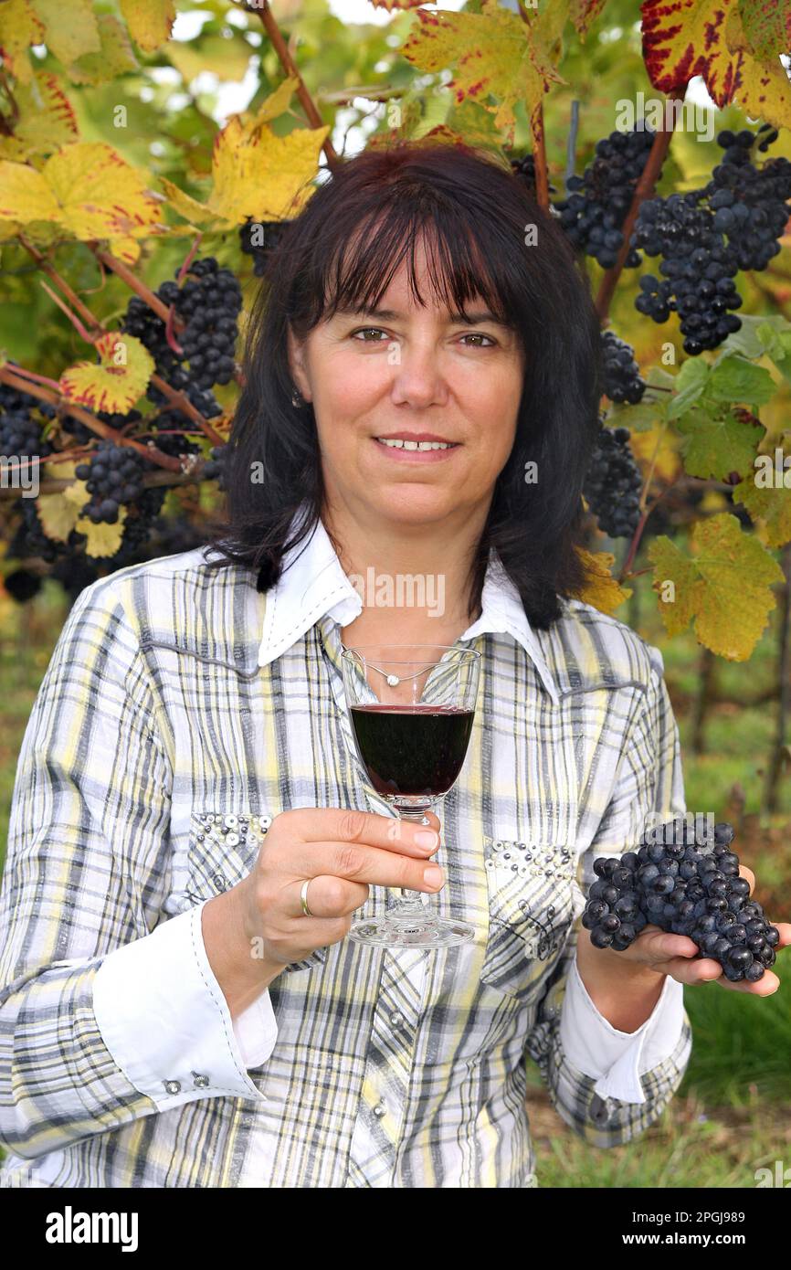 donna con un bicchiere di vino rosso e uva davanti alla vite, mezzo ritratto Foto Stock