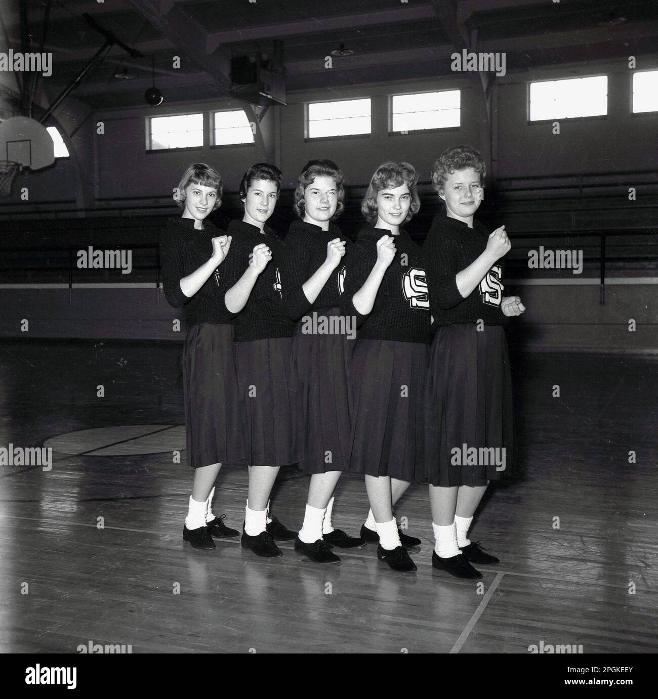 1960s, storico, all'interno di una sala sportiva, una squadra di cheerleaders nei loro abiti; top con loghi, gonne e calzini, in posa insieme per una foto, USA. Squadre di cheerleaders leader spettatori nel tifo sono una tradizione in eventi sportivi americani come il basket e il football americano. Foto Stock