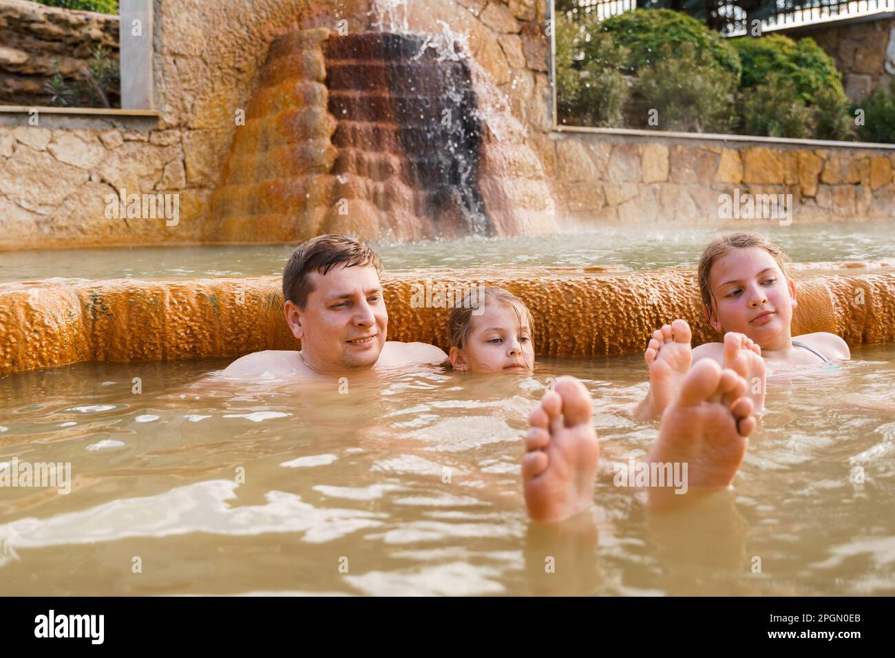 Famiglia, papà e bambini fanno il bagno, nuotano nelle sporche sorgenti termali all'aperto. Piscina con acqua termale e fanghi naturali, argillosi e verdi gialli. Spa wellnes Foto Stock