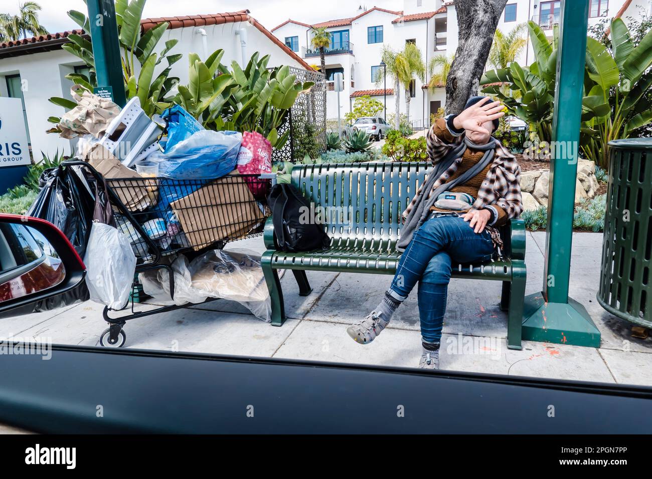 Un maschio senza casa siede su una panchina di fermata dell'autobus con possessi mondani in un carrello di shopping sovraccarico su state Street a Santa Barbara, California. Foto Stock