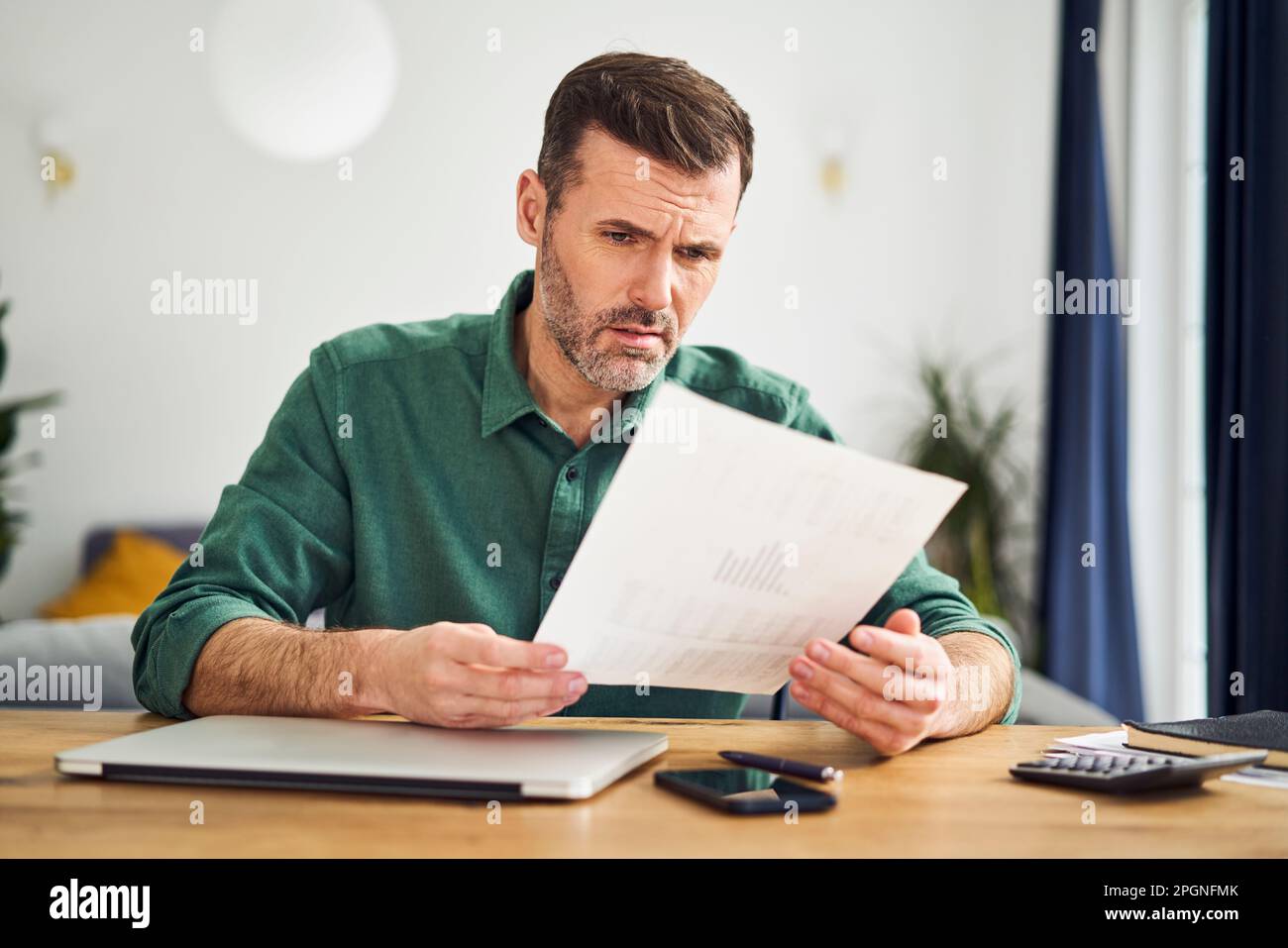 Uomo sconvolto che guarda il documento con figuratori finanziari mentre si siede al tavolo a casa Foto Stock