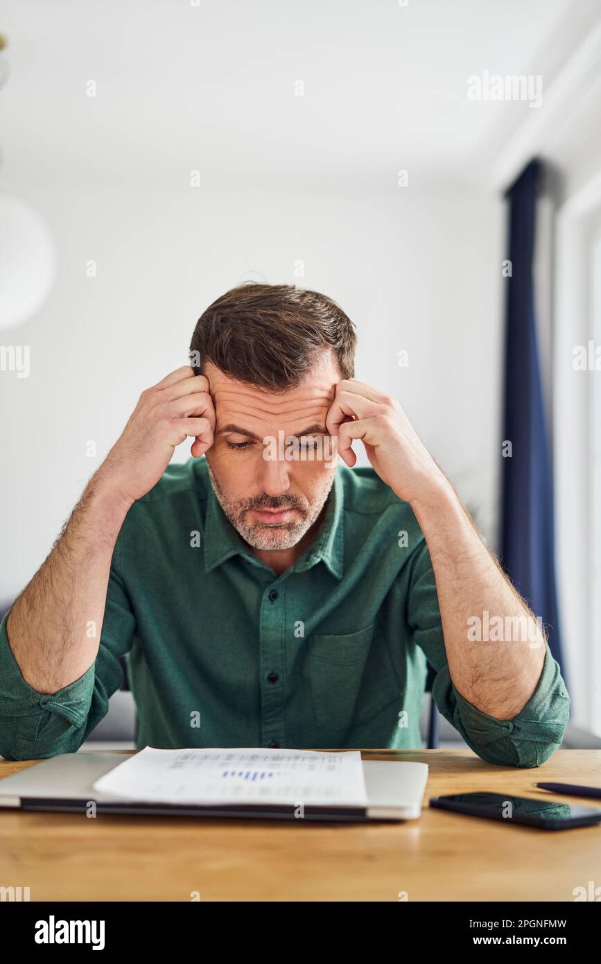 Uomo preoccupato guardando documento con figuratori finanziari mentre seduto al tavolo a casa Foto Stock