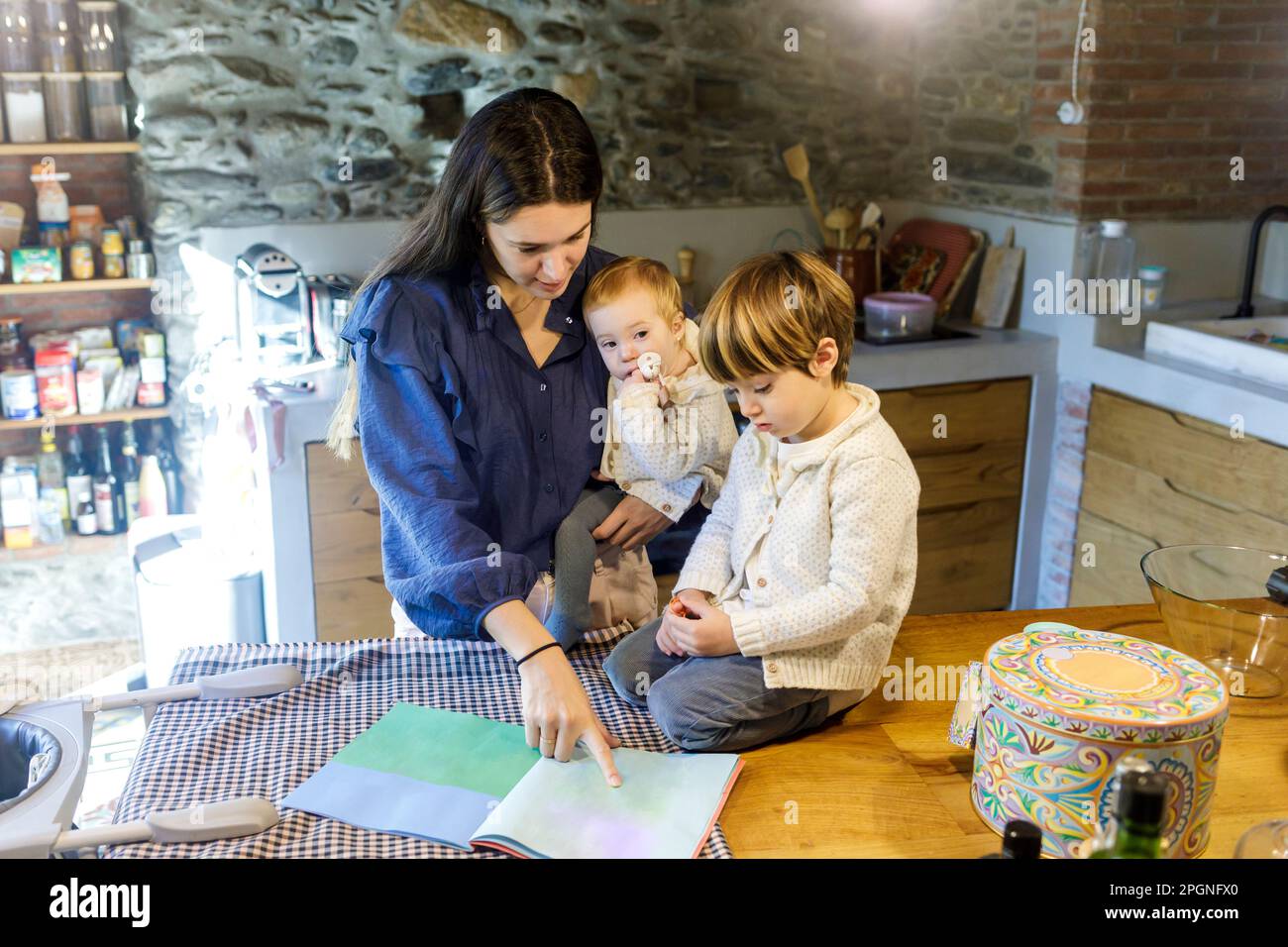 Madre con bambini che legge un libro di immagini in cucina Foto Stock