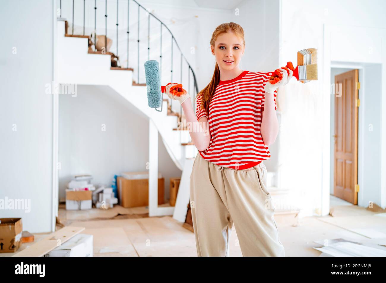 Ragazza giovane sorridente con rullo di vernice e spazzole in piedi a casa Foto Stock