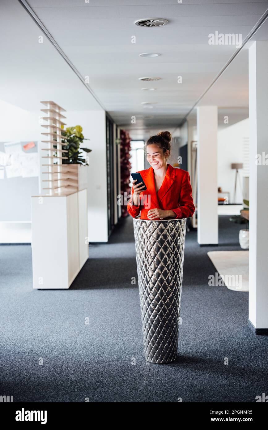 Donna d'affari felice usando il telefono intelligente in piedi all'interno della piantatrice Foto Stock