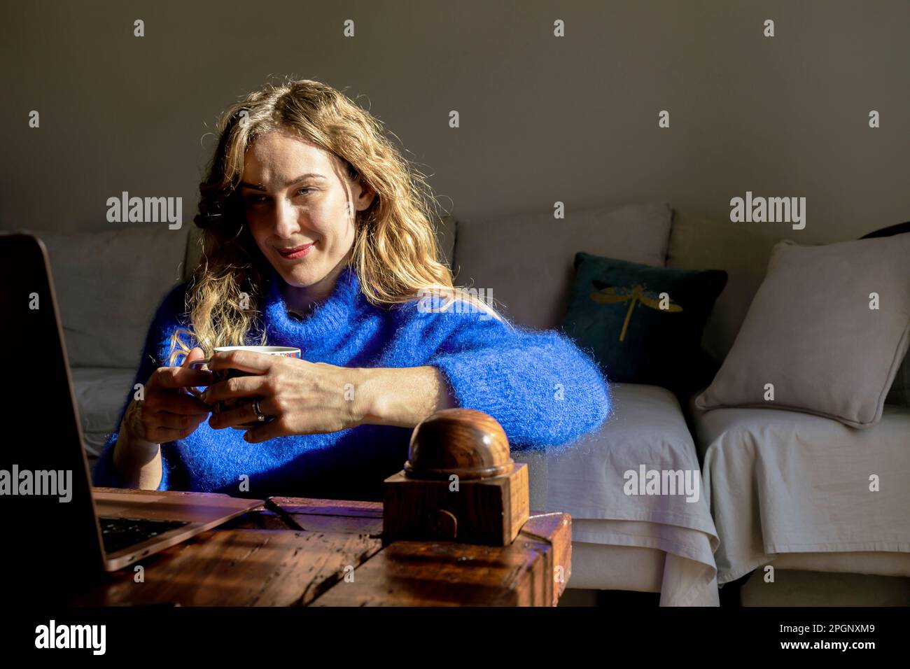 Donna sorridente che tiene una tazza da tè guardando un computer portatile nel soggiorno Foto Stock