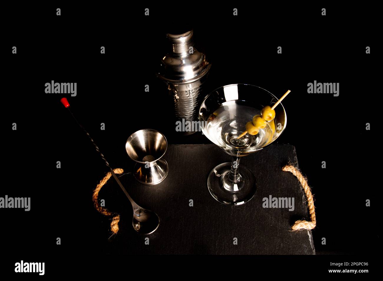 Primo piano di strumenti da cocktail su sfondo nero. Jigger, shaker, cucchiaio e bicchiere martini con tre olive. Vista su Zenith Foto Stock