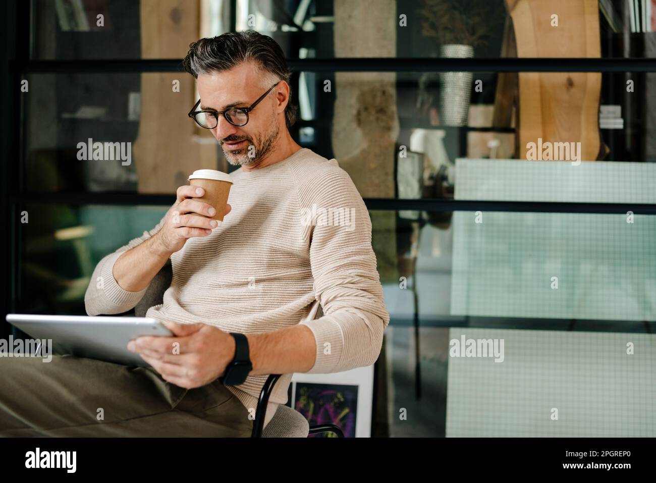 Uomo grigio maturo che beve caffè mentre lavora con un tablet in ufficio Foto Stock