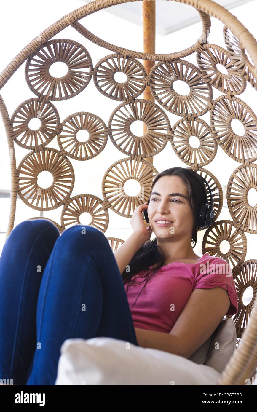 Bella giovane donna caucasica che ascolta musica con le cuffie mentre si siede in altalena a casa. Inalterato, tecnologia, guardare via, stile di vita e rel Foto Stock