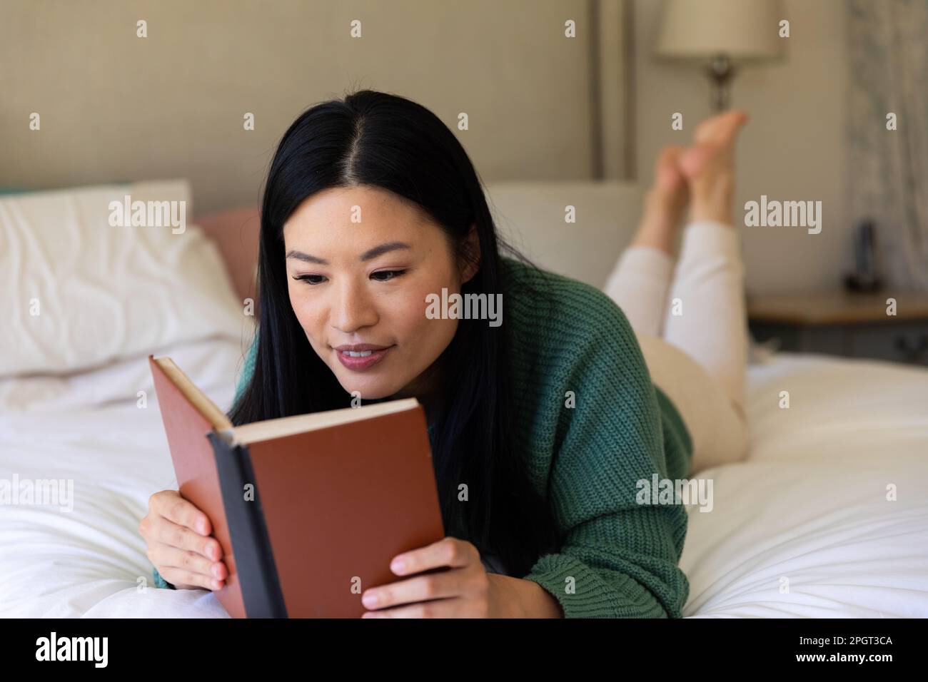 Donna asiatica felice sdraiata sul suo letto leggendo un libro durante il giorno. Stile di vita, tempo libero e relax. Foto Stock