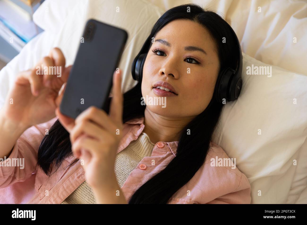 Felice donna asiatica sdraiata sul letto indossando cuffie con smartphone. Stile di vita, tempo libero, comunicazione e relax. Foto Stock