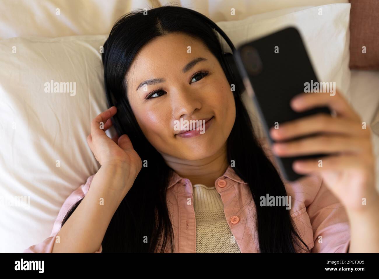 Donna asiatica sorridente sdraiata sul letto indossando le cuffie con lo smartphone. Stile di vita, tempo libero, comunicazione e relax. Foto Stock