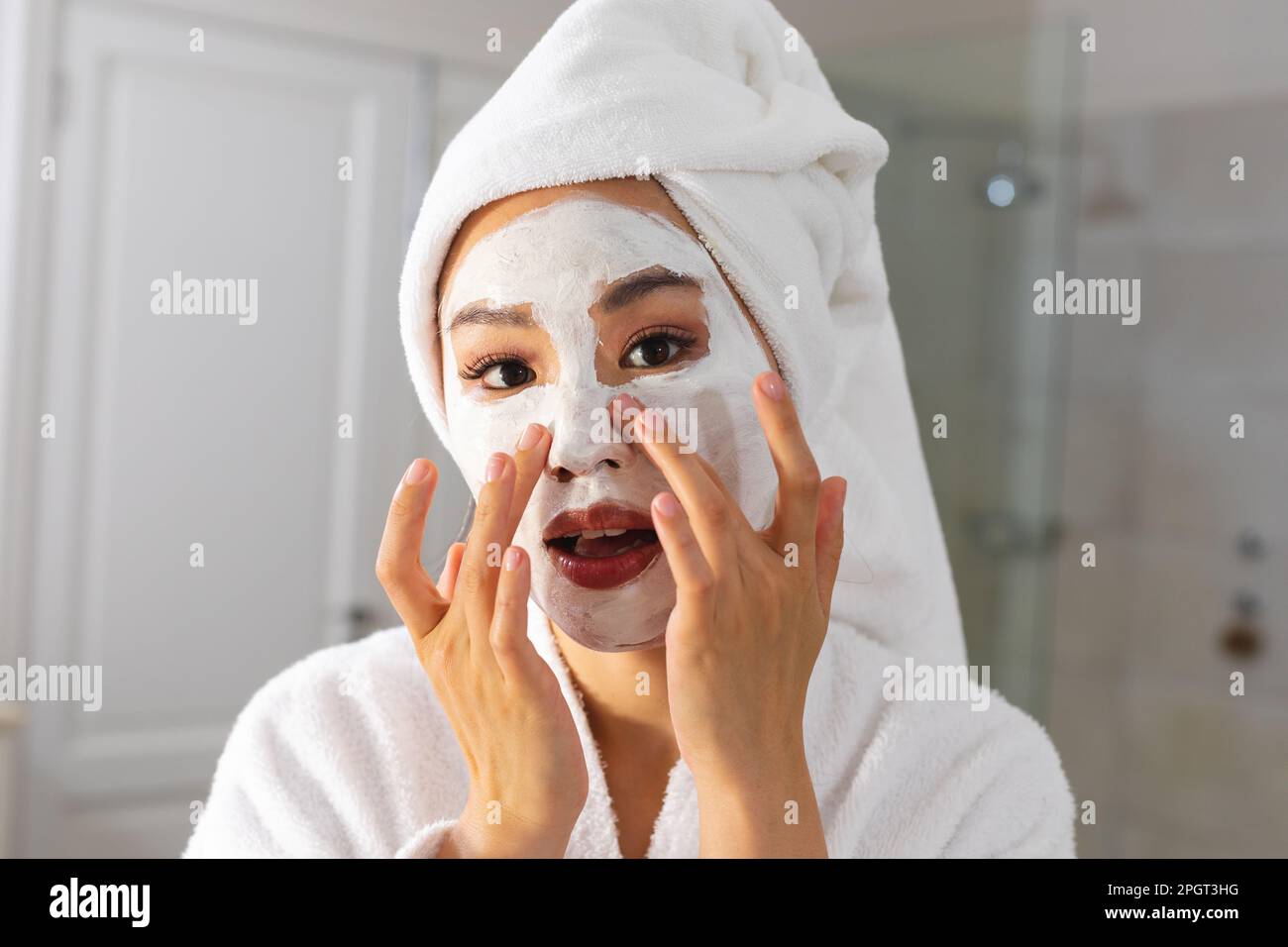 Donna asiatica che indossa accappatoio e asciugamano sulla testa guardando in bagno specchio applicando la maschera di bellezza viso. Stile di vita, cura di sé, salute e bellezza. Foto Stock
