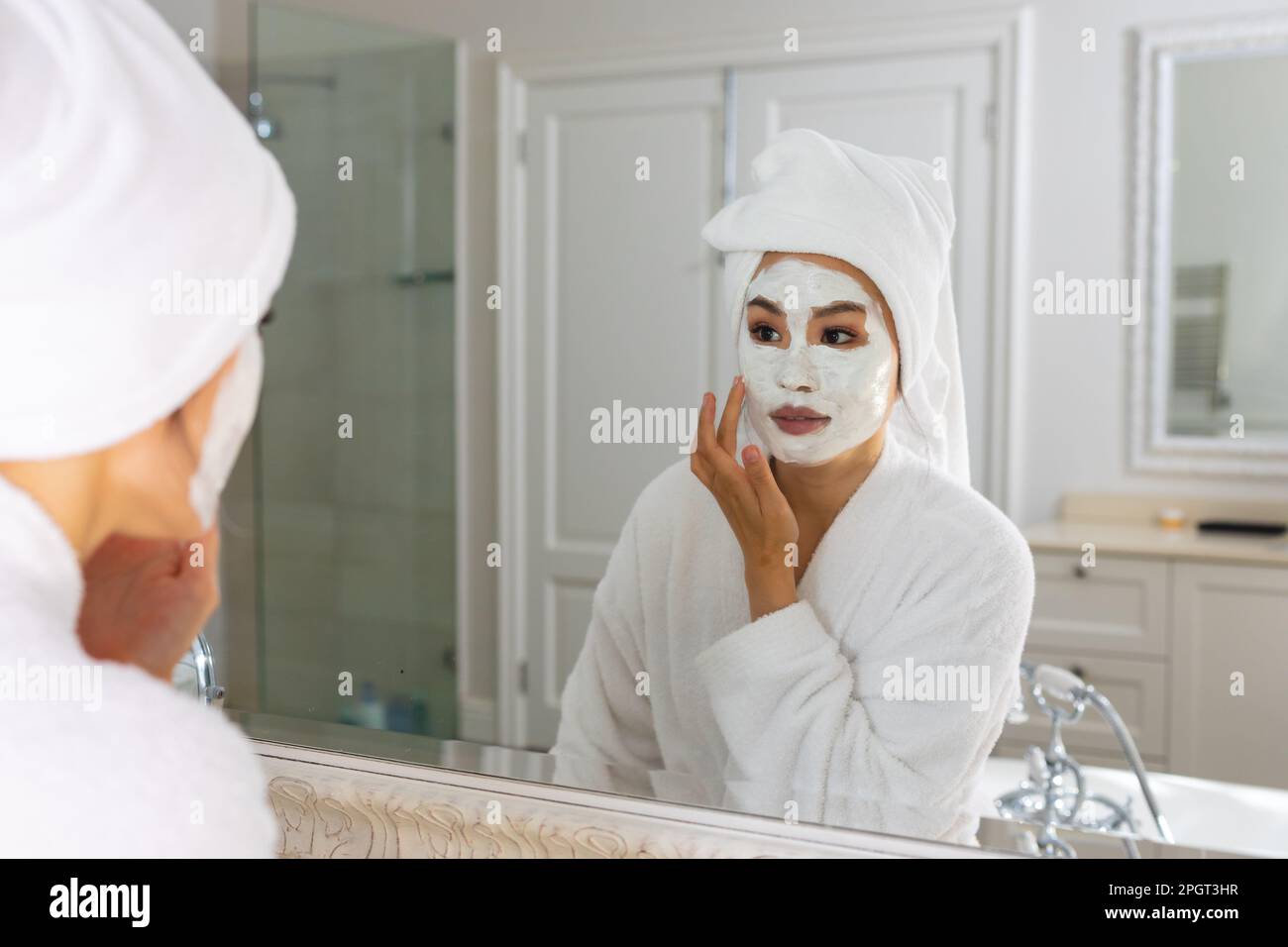 Donna asiatica che indossa accappatoio e asciugamano sulla testa guardando in bagno specchio applicando la maschera di bellezza viso. Stile di vita, cura di sé, salute e bellezza. Foto Stock