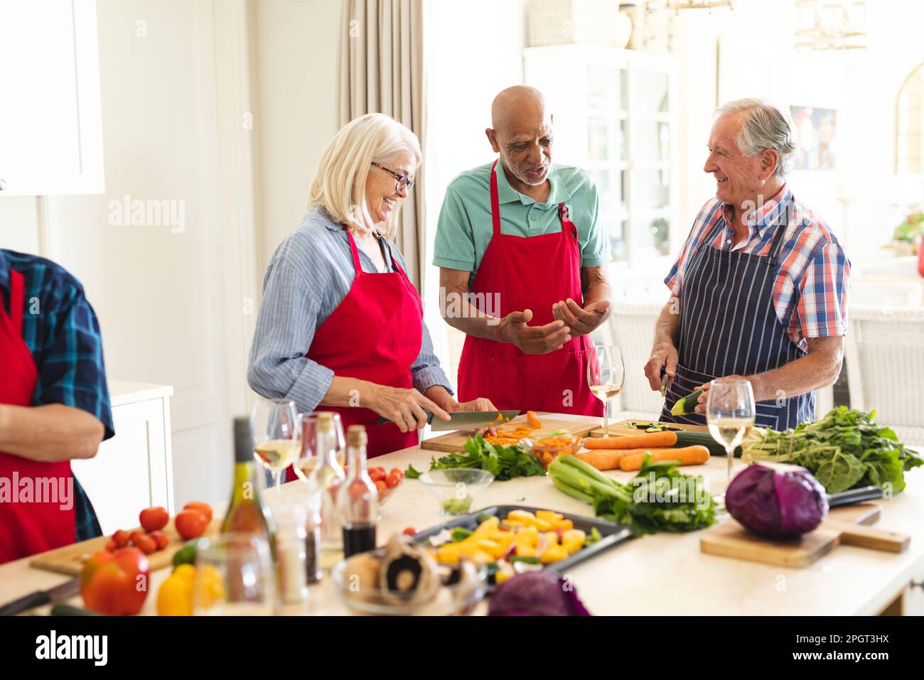 Felice gruppo di amici anziani diversi in grembiuli che parlano durante la lezione di cucina. Pensione, apprendimento, cucina e stile di vita sano. Foto Stock