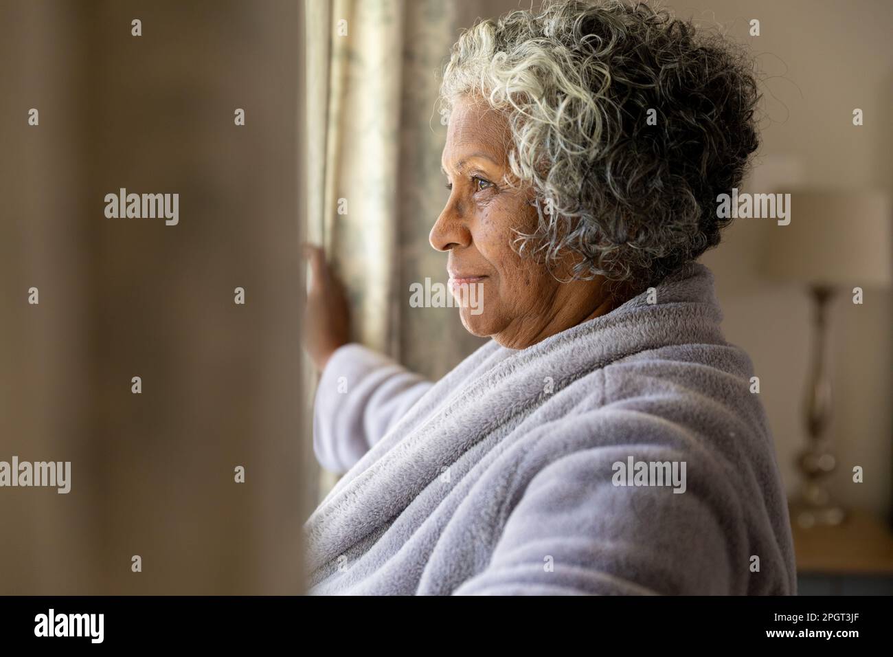 Vista laterale di una donna anziana afroamericana che apre le tende dopo il risveglio al mattino. Copia spazio, inalterato, accappatoio, guardando attraverso la finestra, s Foto Stock