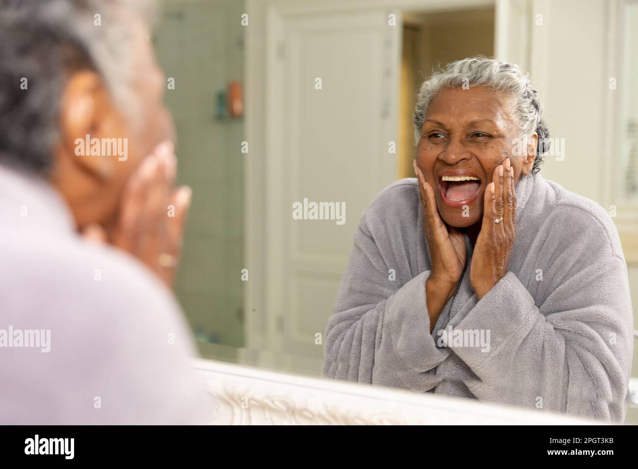 Donna afroamericana anziana che urla allegramente mentre tocca il suo volto e guarda nello specchio. Inalterato, cura del corpo, cura della pelle, invecchiamento, felice, refle Foto Stock