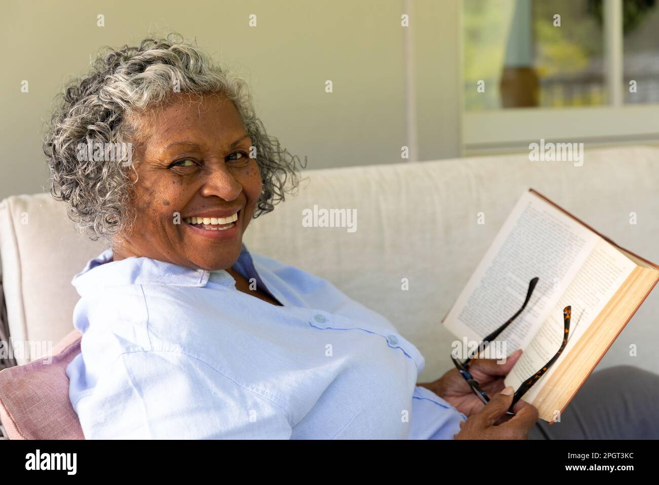 Ritratto di afroamericana senior woman con occhiali e libro sorridente mentre si rilassa sul divano. Copia spazio, inalterato, stile di vita, felice, hobby, hom Foto Stock
