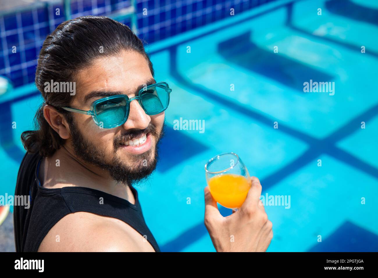 Giovane uomo indiano che indossa occhiali da sole seduti sul bordo della piscina tenendo un bicchiere di succo d'arancia in una calda giornata di sole. Un ragazzo che si rilassa all'aperto vicino alla piscina. S Foto Stock