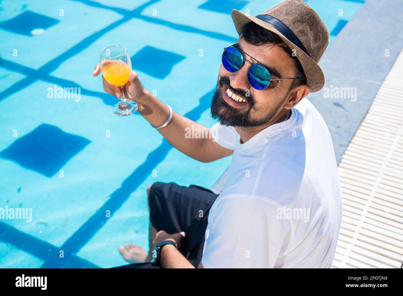 Giovane indiano che indossa cappello e occhiali da sole seduti sul bordo della piscina bere succo d'arancia nella calda giornata di sole. Un ragazzo che si rilassa all'aperto vicino alla piscina. S Foto Stock