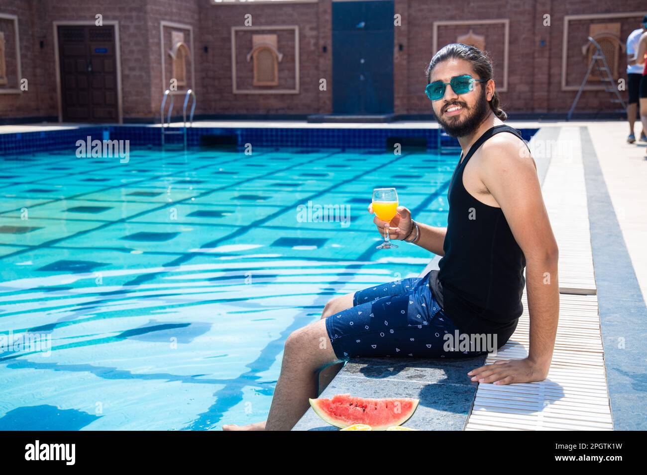 Giovane indiano seduto sul bordo della piscina con i piedi in acqua e tenendo un bicchiere di succo d'arancia in caldo giorno di sole. Ragazzo che si rilassa all'aperto nuotando Foto Stock