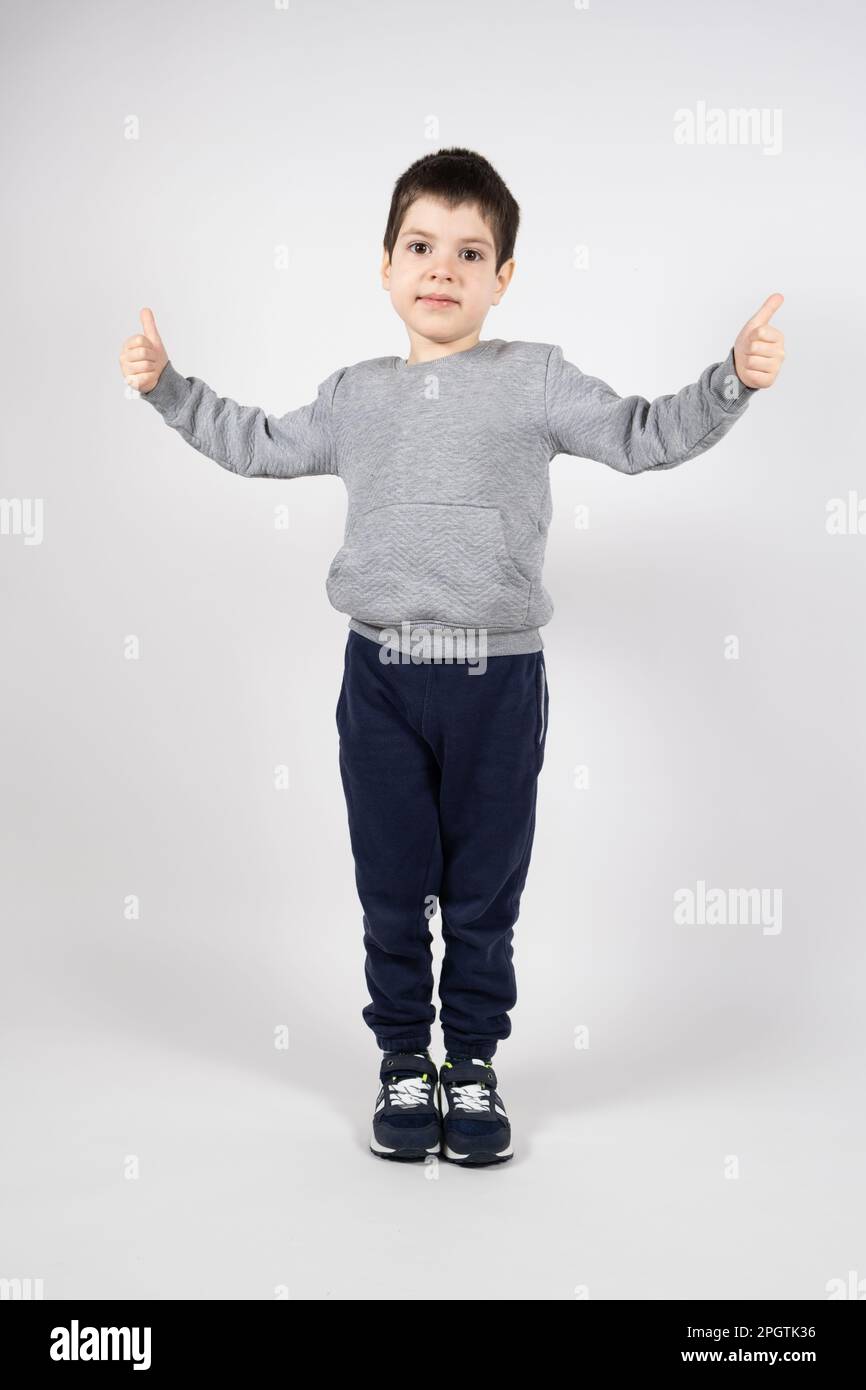 Un ragazzino in una felpa, pantaloni e sneakers che mostra i pollici su uno sfondo bianco. Abbigliamento e calzature per bambini Foto Stock