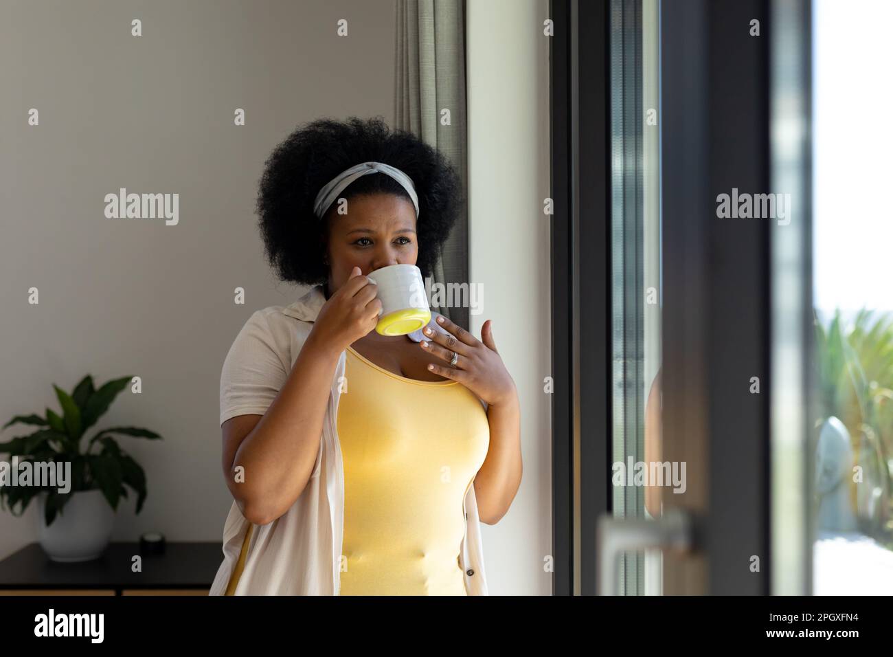 Donna afro-americana premurosa e di grandi dimensioni che guarda fuori dalla finestra bere caffè Foto Stock