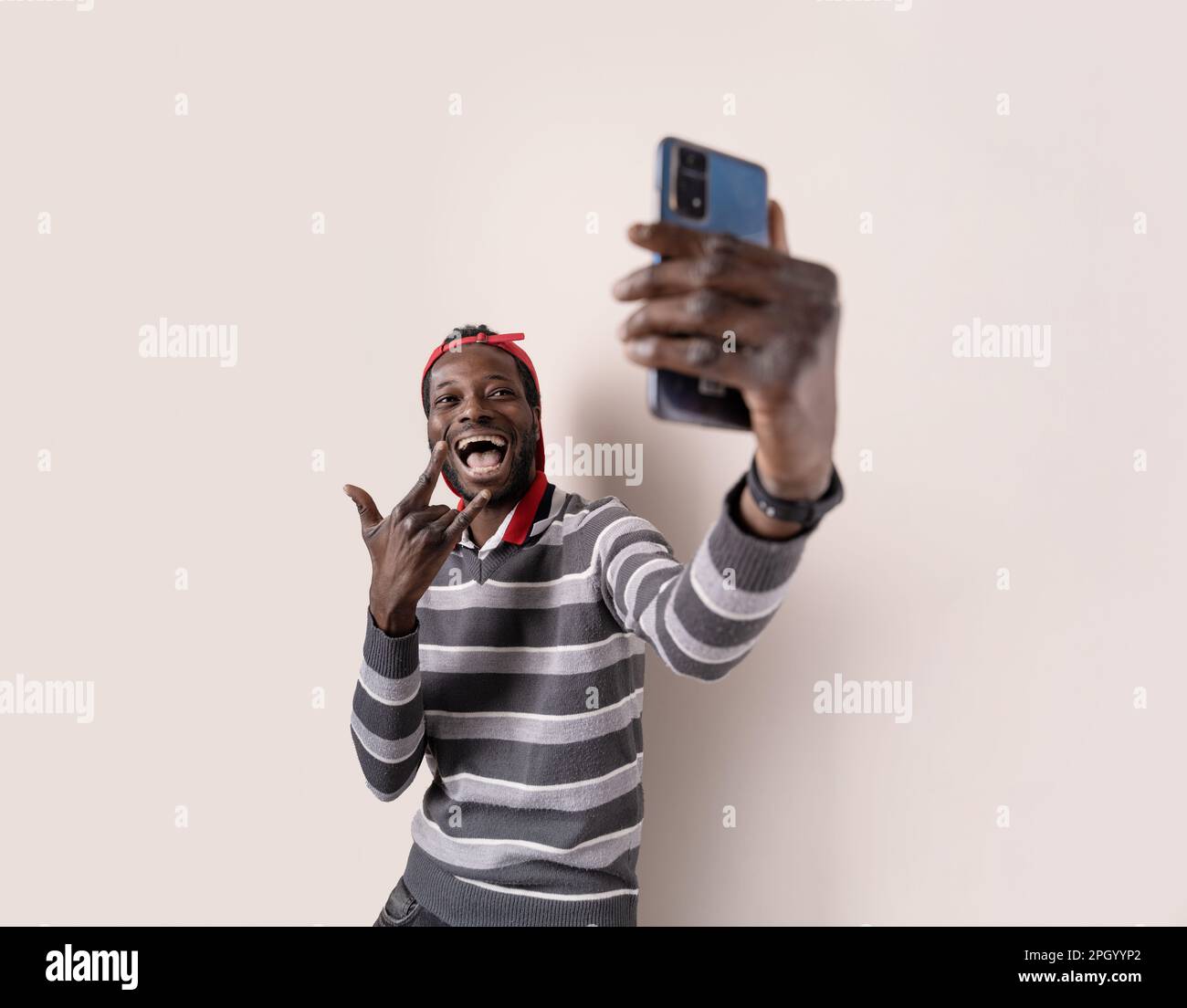 Giovane uomo africano allegro, con un maglione a righe grigie, un volto divertente e un gesto d'amore rock 'n' roll mentre si prende un selfie smartphone. Foto Stock