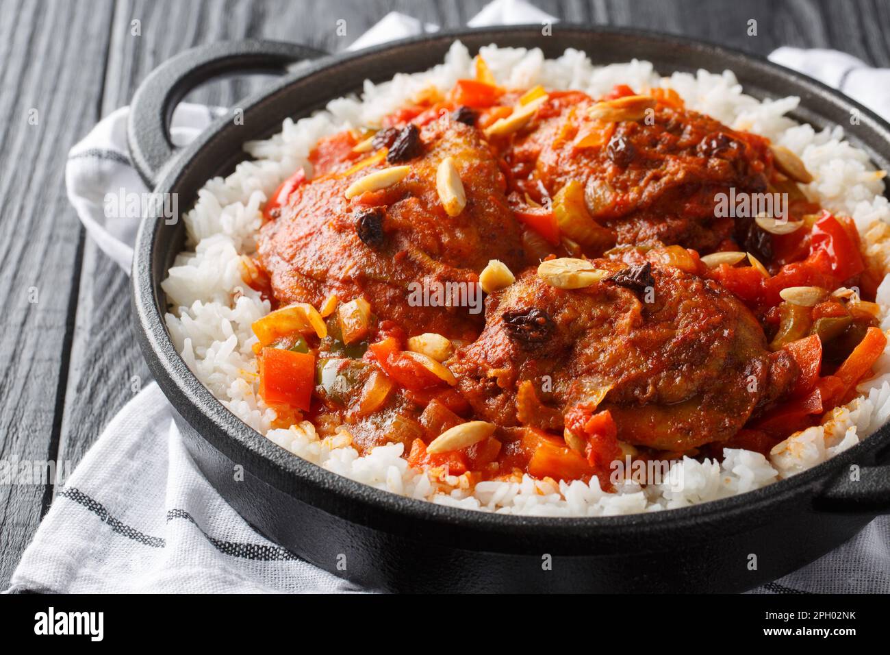 Pollo del capitano fritto e poi stufato con verdure, uvetta, mandorle, salsa di pomodoro e curry, servito con riso in una padella sul tavolo. Orizzontale Foto Stock