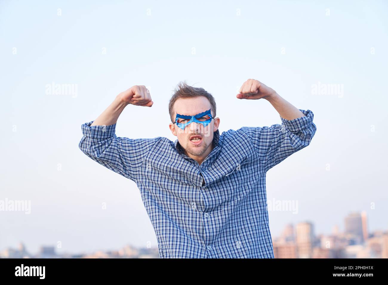 Emozione superpotenza o concetto di pugni pompati. Adorabile uomo caucasico in camicia da notte e maschera blu supereroe con gesto di forza e faccia seria. Immagini di alta qualità Foto Stock