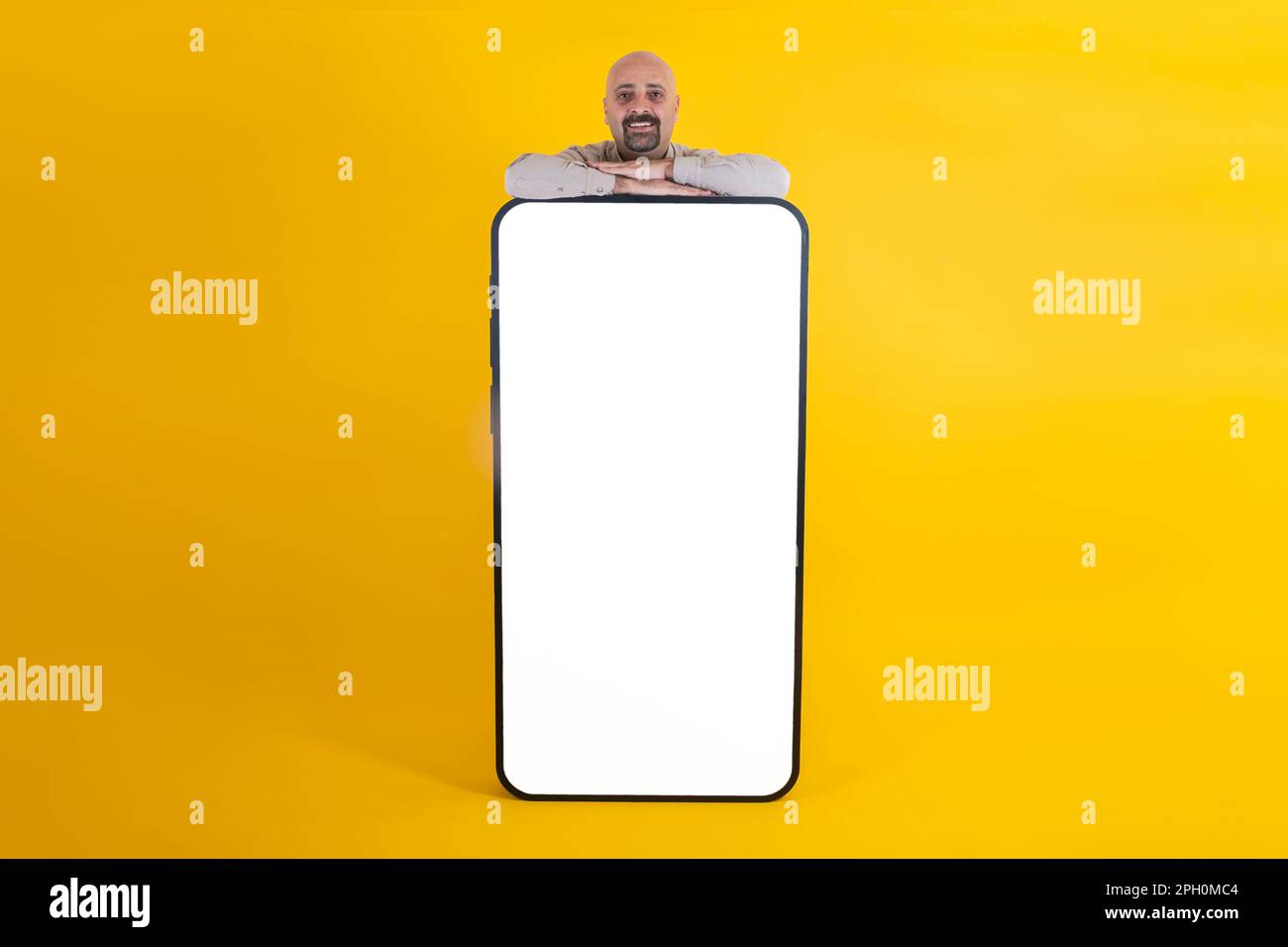 bell'uomo calvo caucasico che si appoggia a un grande smartphone. Spazio copia sfondo giallo studio. Idea di concetto dell'annuncio app. Foto Stock