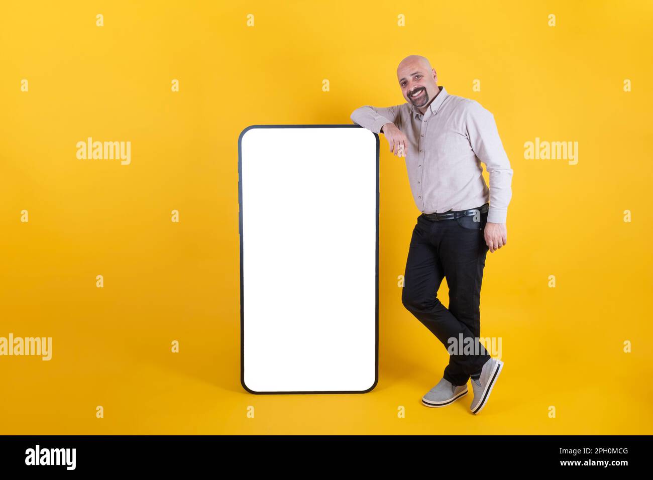 uomo che appoggia un grande smartphone per la pubblicità delle app mobili. Lunghezza completa del corpo di felice uomo sorridente di mezza età vicino a grande schermo vuoto mockup cellulare. Foto Stock