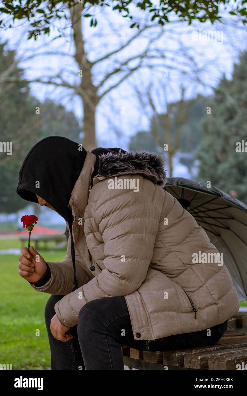 L'uomo con ombrello siede sotto l'albero, tiene il fiore rosso sulla panchina, piovoso Foto Stock