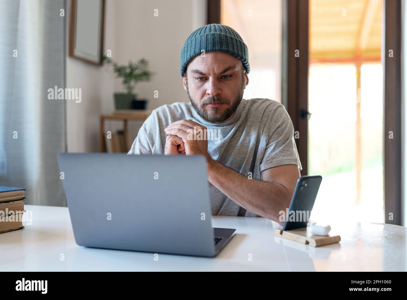 L'uomo in un cappello si siede alla scrivania e guarda una videoconferenza su Internet a casa. Foto Stock