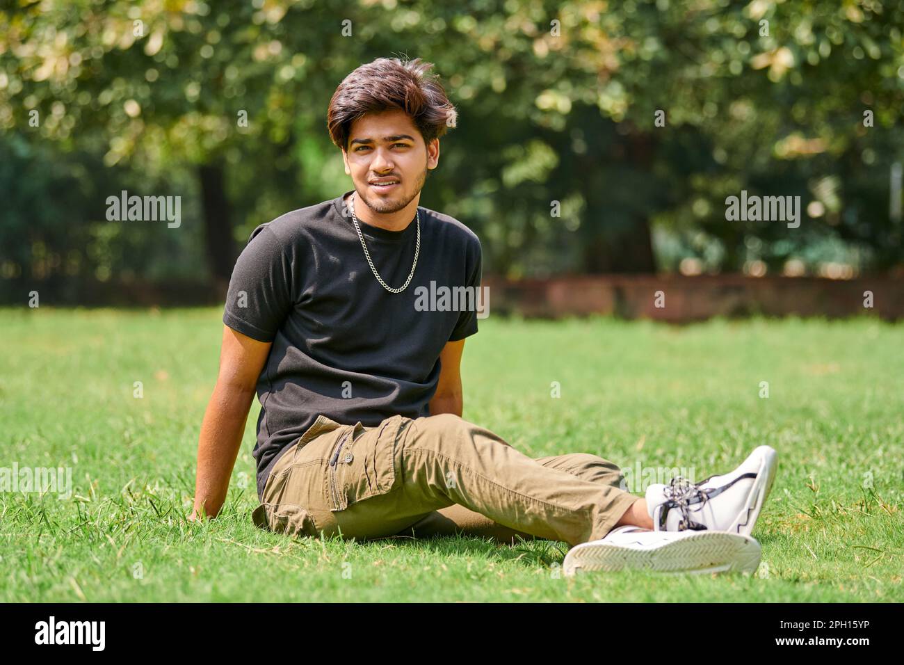 Sorridente ritratto di un uomo indiano in t shirt nera e catena del collo d'argento seduto su prato verde in fondo al parco pubblico, indù attraente portatore maschile Foto Stock
