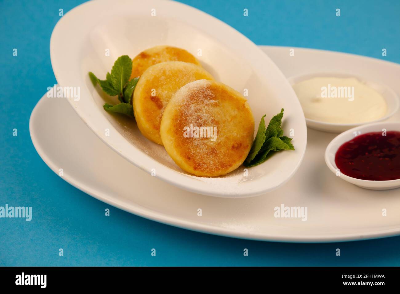 Frittelle di formaggio ricotta rotonda in un piatto bianco su sfondo blu. Cibo prima colazione. Cibo sano. Salsa di panna acida e menta. Primo piano vista dall'alto. Pasto dessert. Frittelle di ricotta al formaggio ricotta. Foto Stock