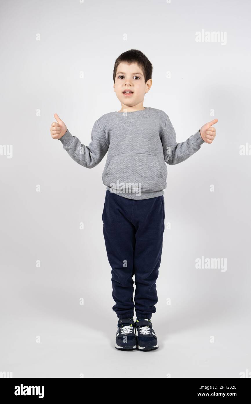 Un ragazzino in una felpa, pantaloni e sneakers che mostra i pollici su uno sfondo bianco. Abbigliamento e calzature per bambini Foto Stock