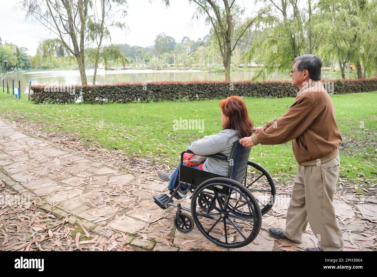 Coppia ispanica anziana passeggiando in un parco, spinge la sua sedia a rotelle lungo un sentiero. Foto Stock