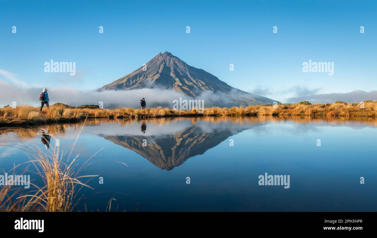 Due persone trekking circuito Pouakai. Il Monte Taranaki si rifletteva nel tarn al sole della mattina presto. Nuova Zelanda. Foto Stock