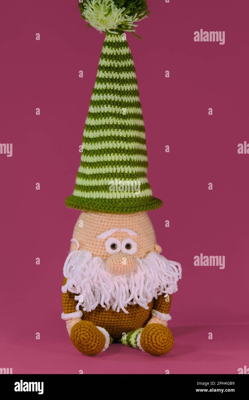 Amigurumi una nana con cappello a strisce verdi e barba bianca su sfondo rosa. Soffice giocattolo fai da te in cotone naturale e lana. Vecchio accovacciato, handma Foto Stock
