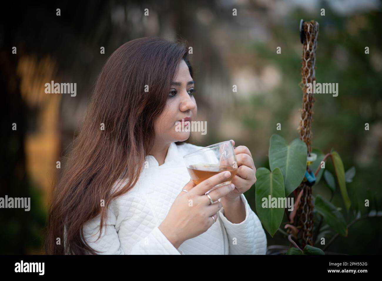 Giovani donne in possesso di una tazza da tè trasparente con entrambe le mani e tè nero in un giardino con giacca di lana bianca Foto Stock