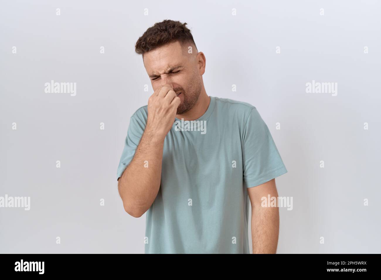 Uomo ispanico con barba in piedi su sfondo bianco odore qualcosa di puzzolente e disgustoso, odore intollerabile, tenendo il respiro con le dita sul nos Foto Stock