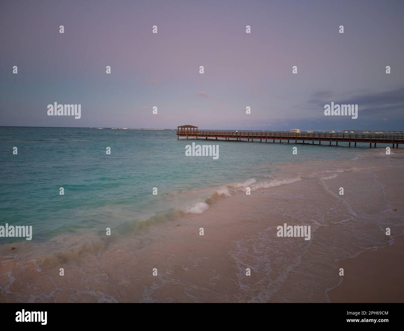 Molo in legno sul mare e spiaggia sabbiosa. In lontananza si può vedere la città sulla costa. Onde marine leggere corrono verso la riva. Crepuscolo. Bellezza di natu Foto Stock